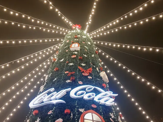 The image shows a large Christmas tree decorated with lights and ornaments, with the iconic Coca...