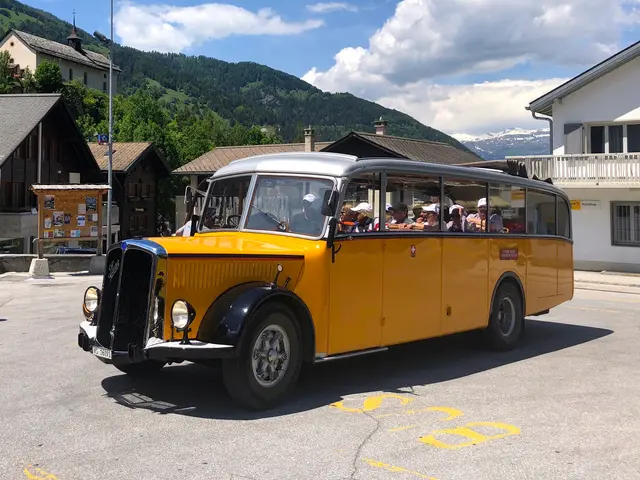 The image shows a yellow bus driving down a street lined with houses and trees, with mountains in...