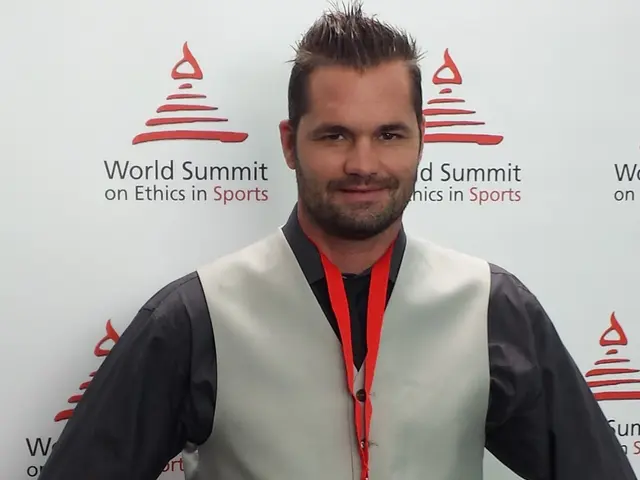 The image shows a man in a suit standing in front of a wall with a banner behind him that reads...
