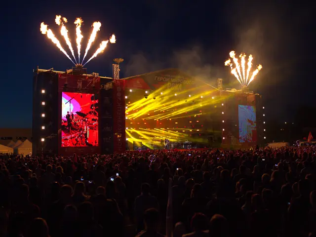 The image shows a large crowd of people gathered at a music festival, with a stage in the center of...