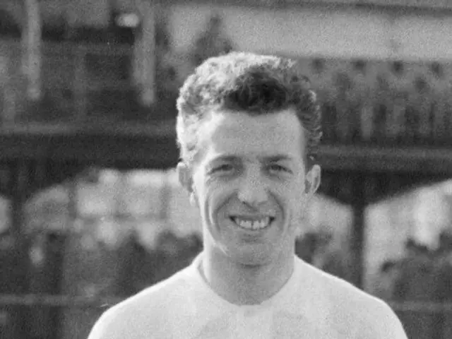 The image shows a black and white photo of a man standing on a soccer field, smiling. He is wearing...