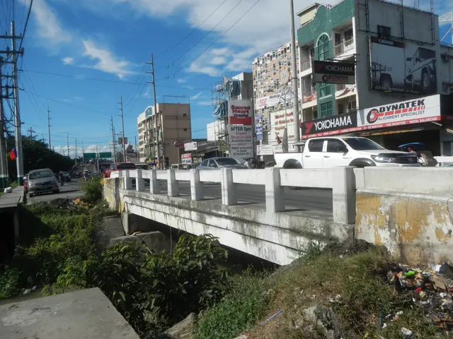 The image shows a city street with cars driving on it, a bridge over a river, buildings with...