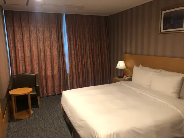 The image shows a hotel room with a large bed covered in a blanket and pillows, a chair, a table, a...