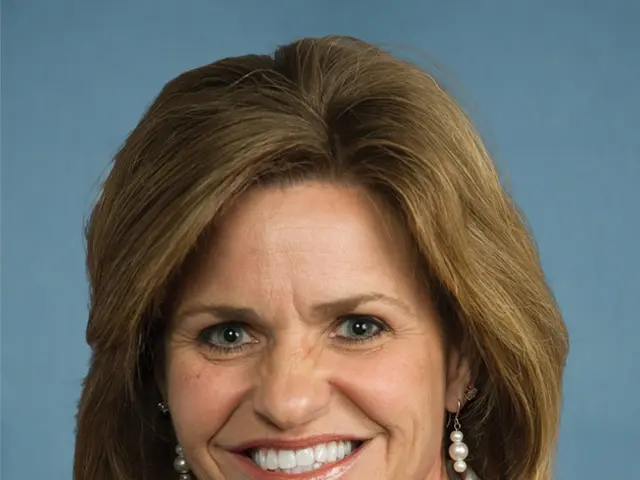 The image shows a woman in a business suit smiling at the camera against a blue background. She...