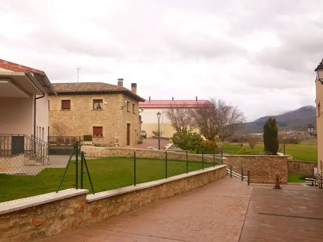 The image shows a building with windows, railings, steps and a roof, surrounded by a metal fence,...