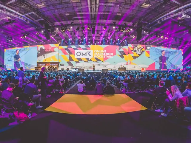 The image shows a large crowd of people sitting in chairs in front of a stage, illuminated by...
