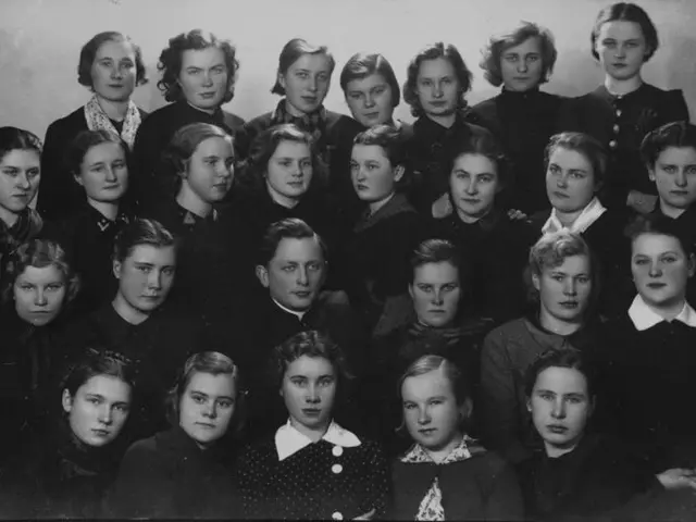 The image shows a black and white photo of a group of women in school uniforms, with a wall in the...