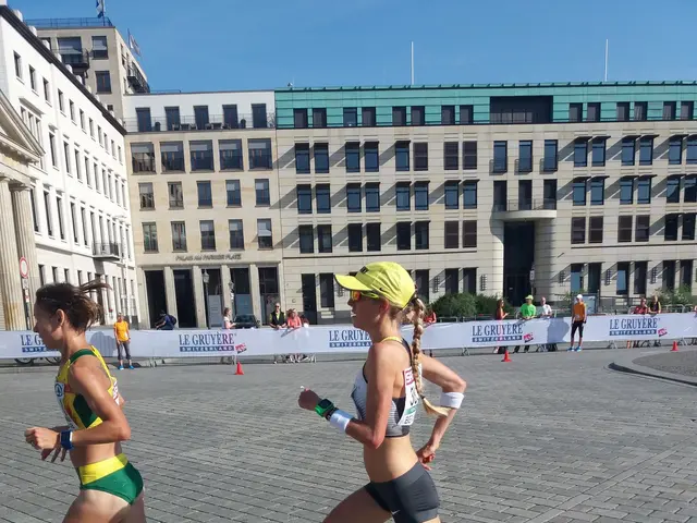 The image shows two women running in a marathon on a city street, surrounded by a group of people...