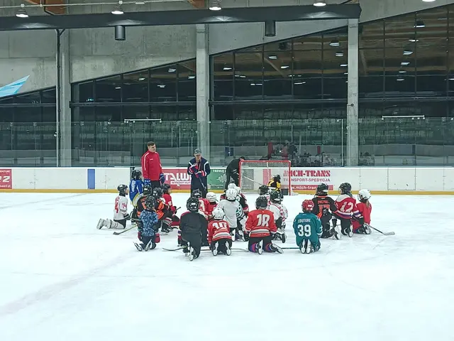 The image shows a group of children sitting on top of an ice rink, wearing helmets and holding...