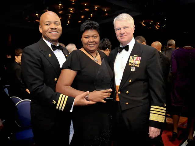 The image shows two men and a woman standing next to each other on a red carpet. The woman is...