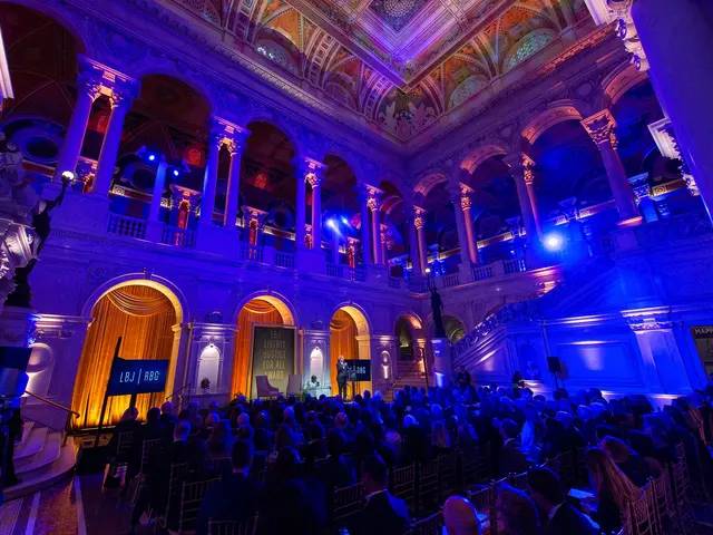 The image shows a large group of people sitting in chairs in a large room with a stage in the...
