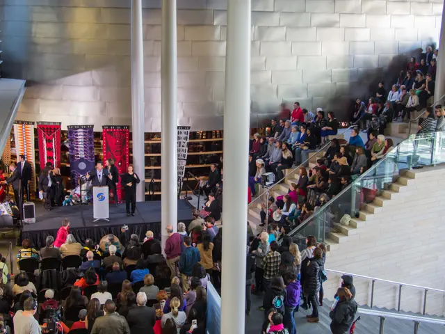 The image shows a large group of people standing in front of a crowd of people sitting on chairs,...