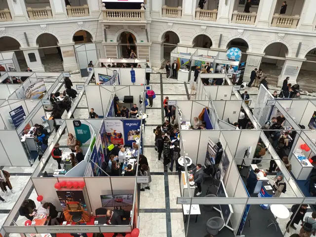 The image shows a large group of people standing around a room filled with booths, banners, tables,...