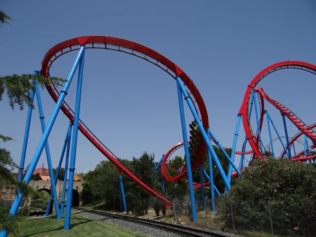 The image shows a roller coaster in the middle of a park, surrounded by lush green grass, trees,...