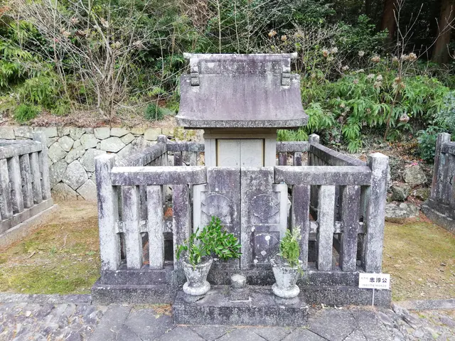 The image shows a small shrine in the middle of a forest, surrounded by a wooden fence and a stone...