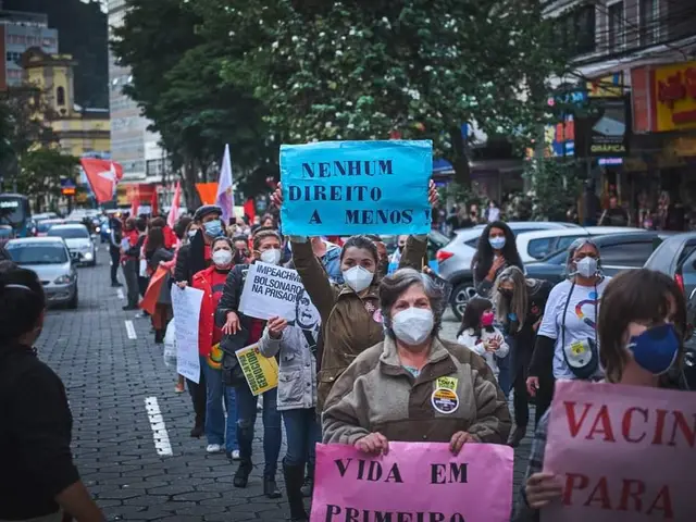 The image shows a group of people wearing masks and holding signs in the middle of a street,...