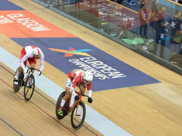 The image shows two men wearing helmets and riding bicycles on a track in front of a crowd. On the...