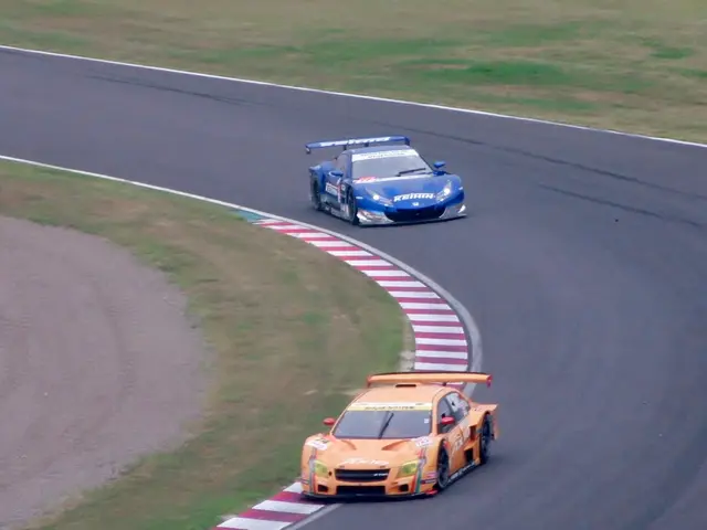 The image shows two sports cars racing on a race track, with one car in orange and the other in...