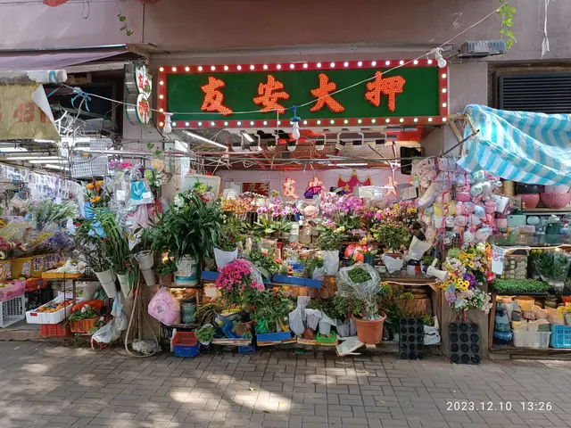 The image shows a bustling flower market in Hong Kong, with a variety of colorful flowers, plants,...