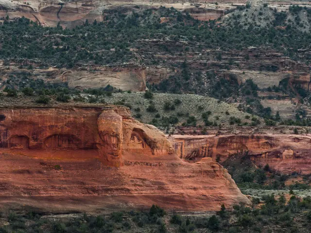 The image shows a large rock formation in the middle of a desert, surrounded by trees and hills. At...
