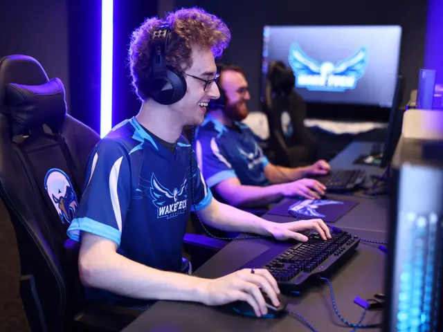 The image shows two men sitting at a desk in front of a computer, wearing headsets and smiling. On...