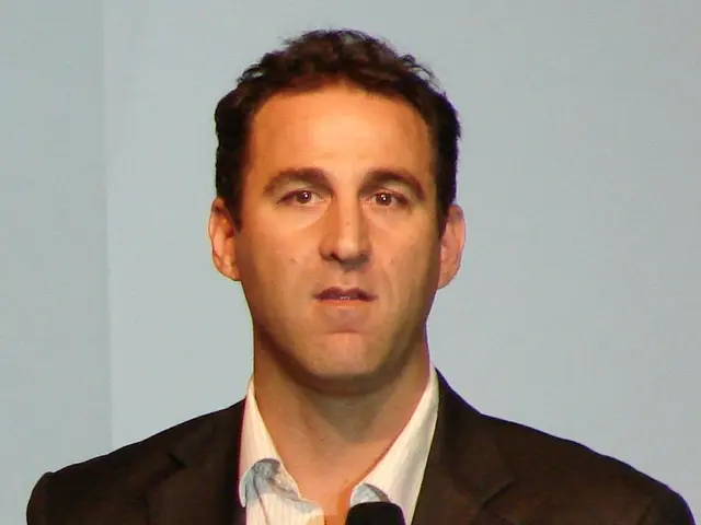 The image shows a man in a suit holding a microphone, standing in front of a screen. He has a...