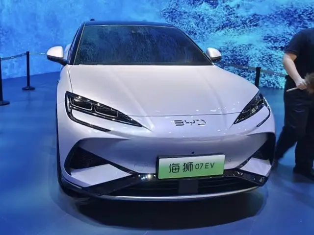 The image shows a white car on display at an auto show, with a person standing to the right of it...