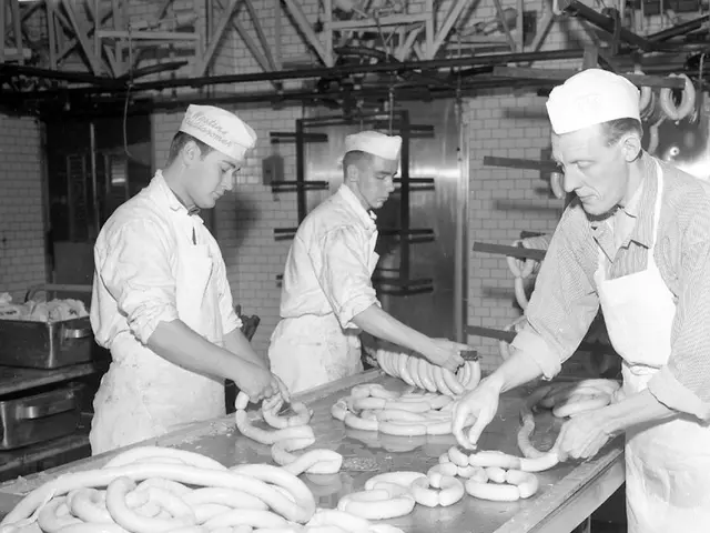 The image shows three men wearing aprons and caps standing in a factory, preparing sausages on a...