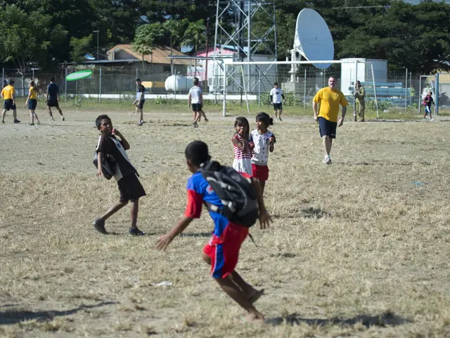 The image shows a group of children playing soccer on a field surrounded by a metal fence, a dish...