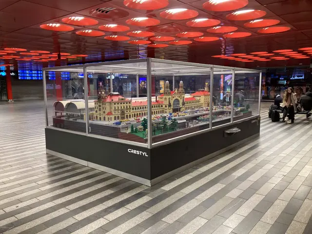 The image shows a display case filled with lots of Legos in an airport, with people sitting on...