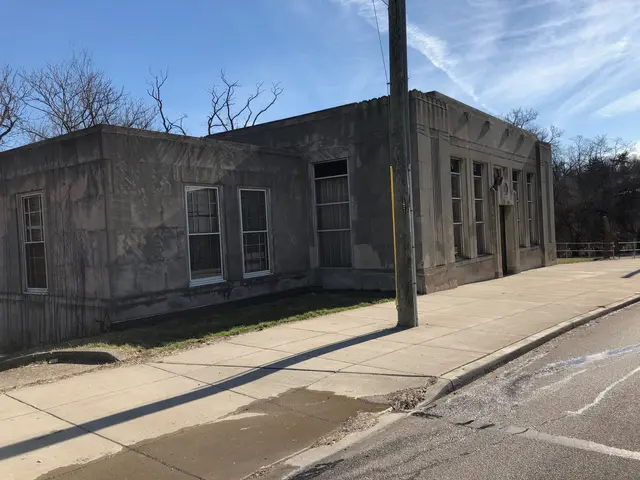 The image shows an old building on the corner of a street, with windows, a pole with wires, grass,...