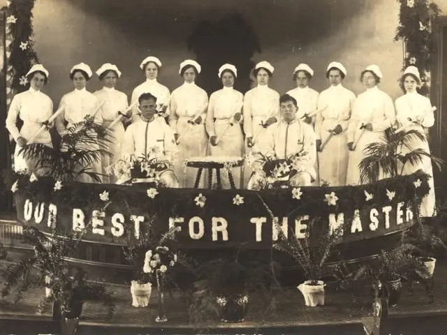 The image shows a group of women standing on top of a stage, wearing white dresses and caps. In the...