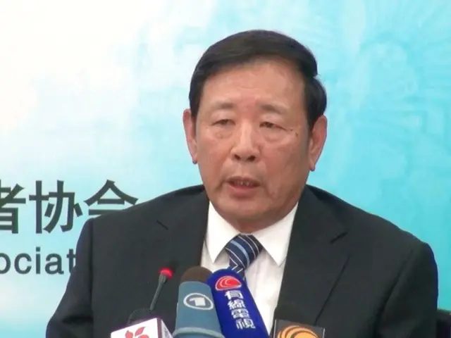 The image shows a man in a suit and tie sitting at a table with microphones in front of him and a...