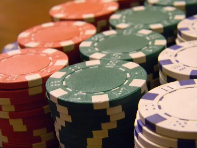The image shows a pile of poker chips of various colors sitting on top of a wooden table. The chips...