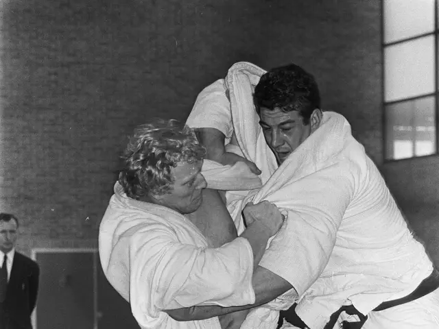 The image shows two men in judo uniforms engaged in a fight in a gym, with a person standing in the...
