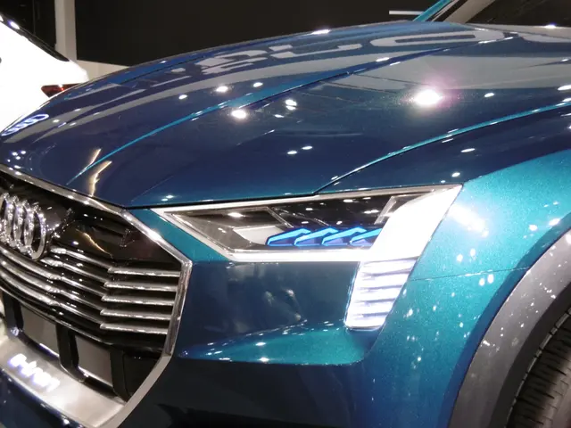 The image shows a blue Audi e-tron concept car on display at an auto show, with a white car in the...