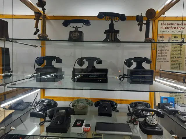 The image shows a display case filled with lots of different types of telephones on glass shelves,...