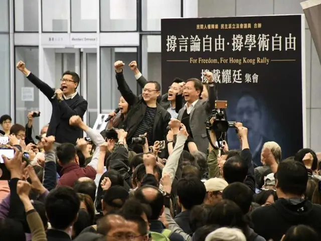 The image shows a group of people standing in front of a crowd of people, some of whom are holding...