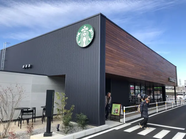 The image shows a Starbucks coffee shop on the corner of a street, with two people walking on the...