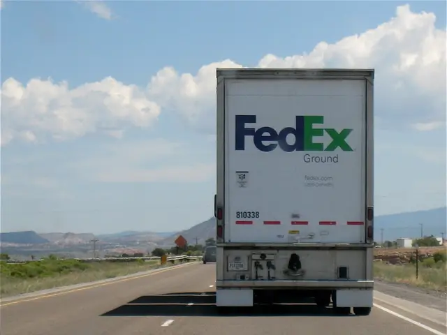 The image shows a FedEx truck driving down a highway with mountains in the background. The truck is...