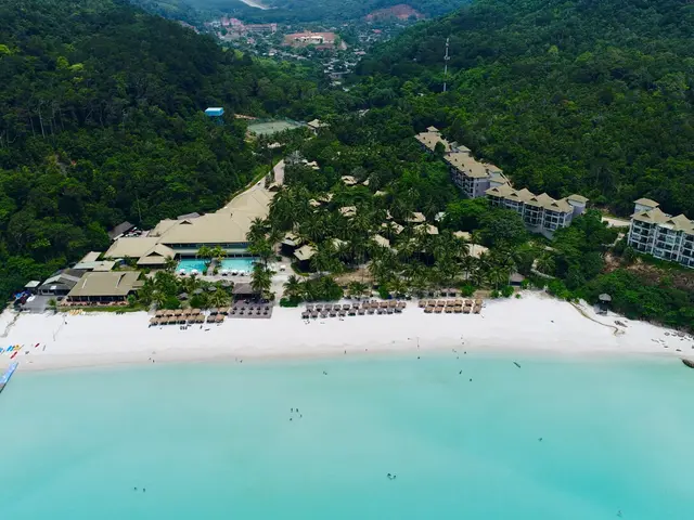 The image shows an aerial view of a resort on the beach, with a bridge on the left side, buildings,...
