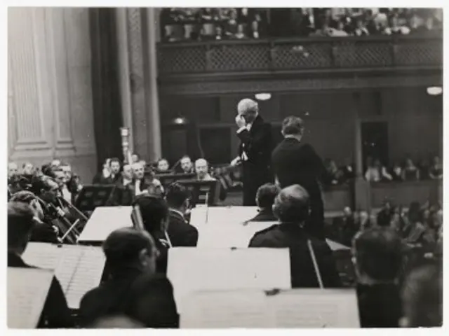 The image shows a black and white photo of a conductor standing in front of an orchestra, with a...
