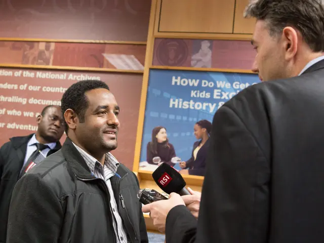 The image shows a man in a suit talking to a reporter in front of a wall adorned with banners. The...