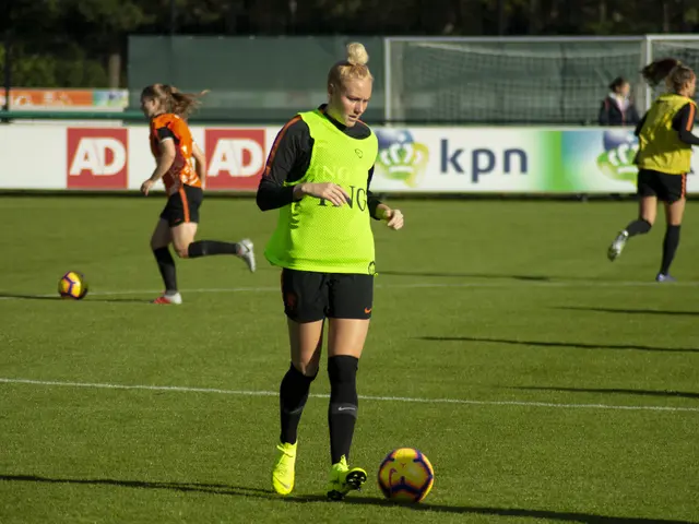 The image shows a group of women playing soccer on a field surrounded by trees. There are banners...