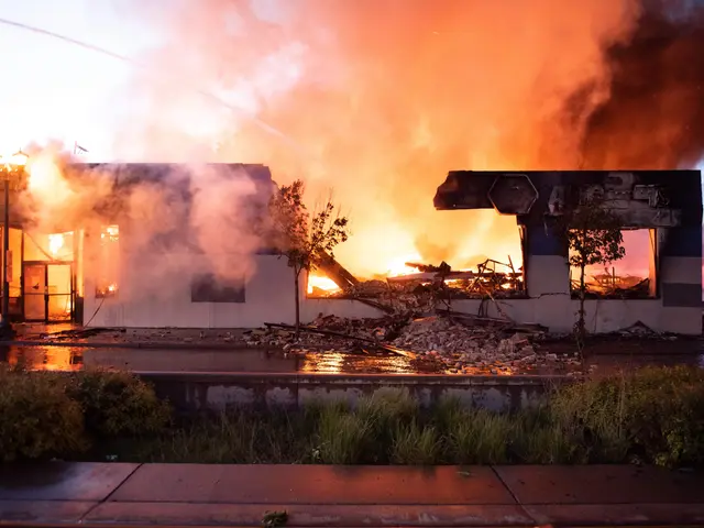 The image shows a house engulfed in flames on the corner of a street, with smoke billowing up into...