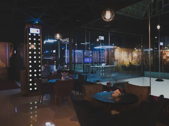 The image shows a restaurant with tables and chairs in front of a large window, illuminated by...