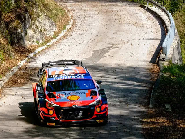 The image shows a rally car driving down a winding road surrounded by trees and plants on either...
