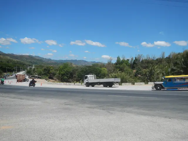 The image shows a truck driving down a street lined with traffic cones, with people riding...