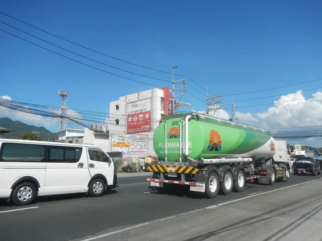 The image shows a large tanker truck driving down a street next to a white van. In the background,...