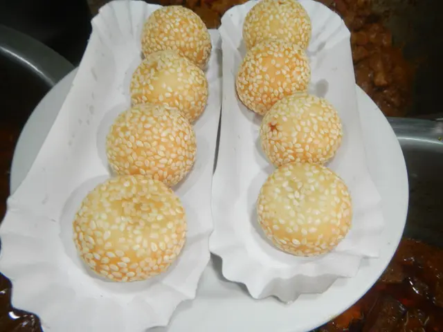 The image shows a white plate topped with six sesame buns on top of a table, with a bowl of food in...